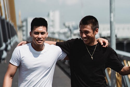 Young happy ethnic male friends embracing while spending time together and talking on bridge in city