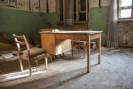 一间尘土飞扬的废弃客房里，柔和的自然光线下摆放着一张旧木制书桌和椅子
