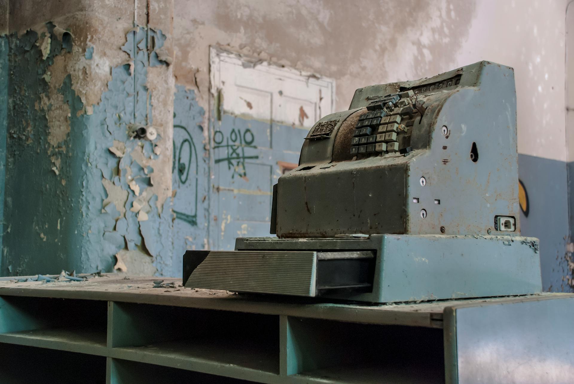 Old cash register sitting in a decaying, abandoned building with peeling paint and debris.