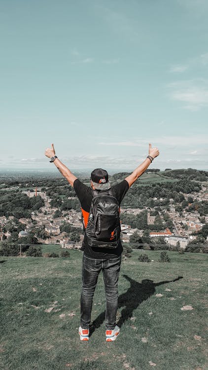 Back view of unrecognizable young male traveler in trendy outfit showing thumbs up gesture while admiring nature standing on green hill on sunny day