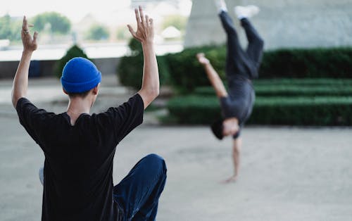 Back view of faceless male in casual clothes instructing friend while performing handstand on city street