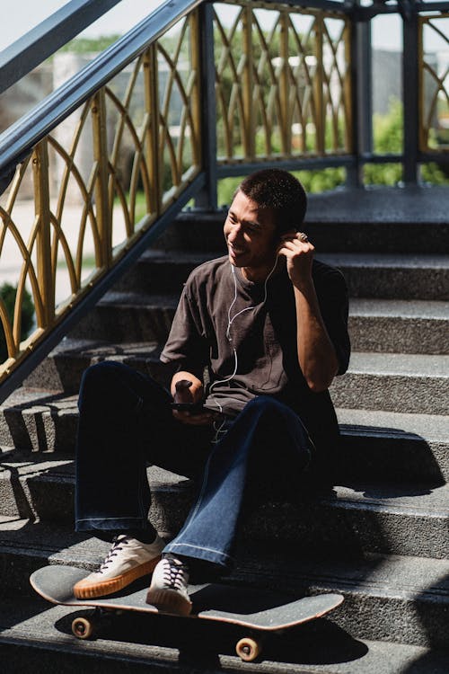 Full body of Asian male in casual clothes sitting on stairs with skateboard and listen to music with earphones