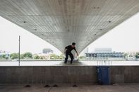 在城市的海边，一名无法辨认的男性在训练期间全身骑在滑板上，位于海桥下的栏杆附近