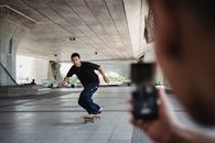 Full body of sportive man riding on skateboard under bridge and looking at camera of smartphone during training in street