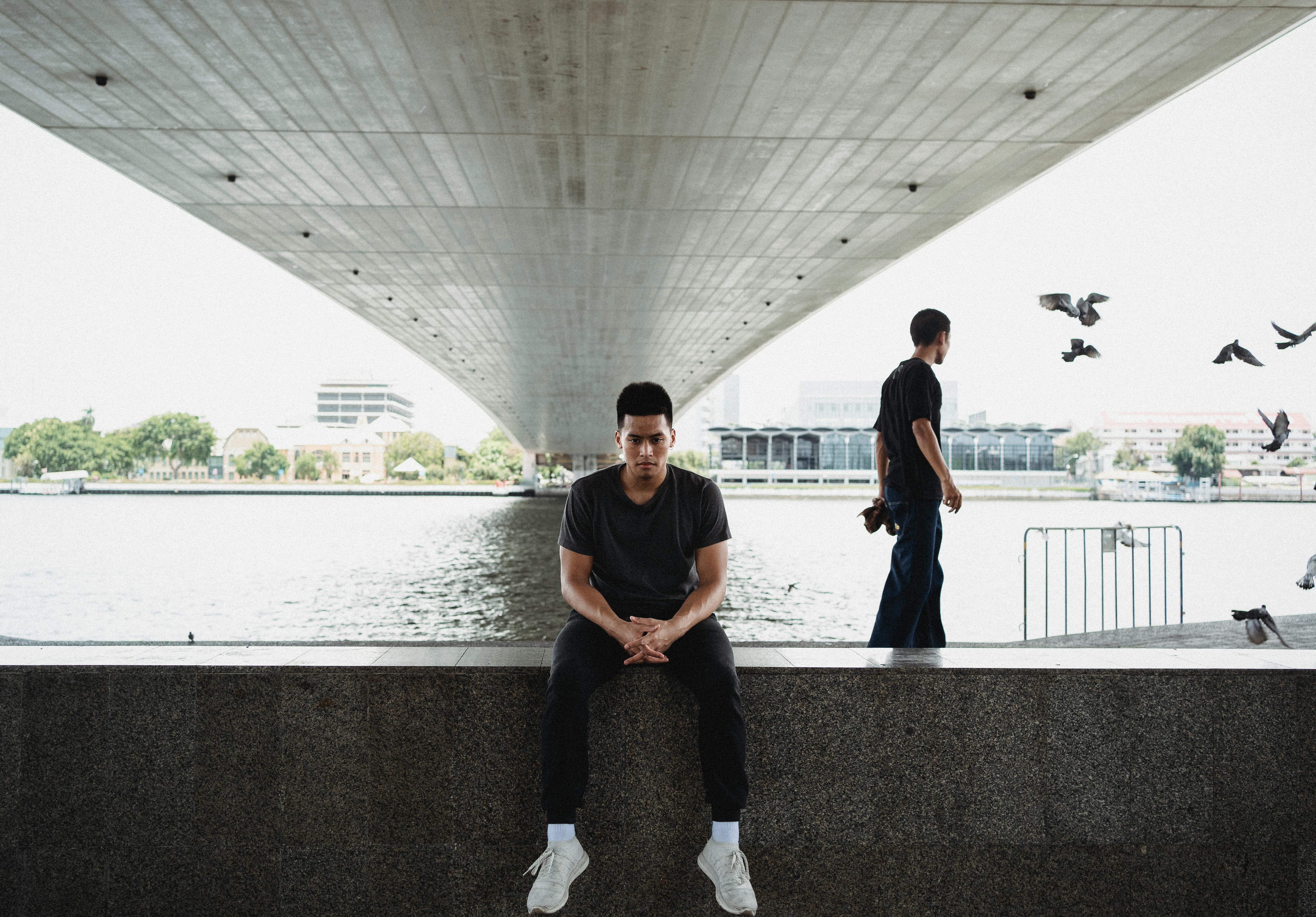 一个男人坐在桥下，河边，周围有鸟类和现代建筑