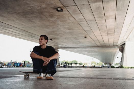 城市桥下，一位亚洲男性滑板运动员静坐，描绘出一种宁静而独立的氛围