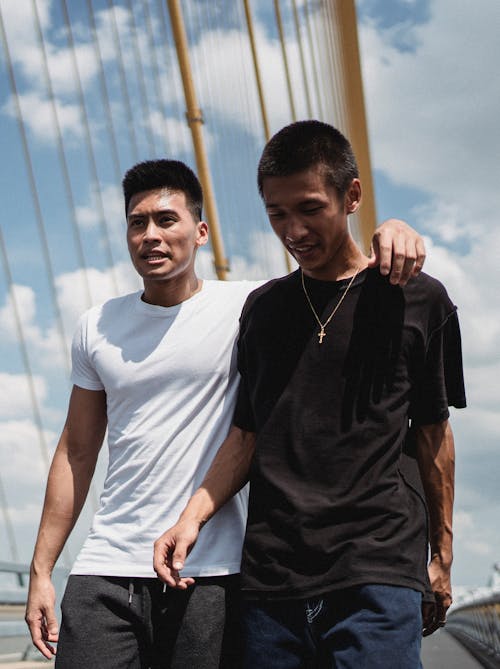 From below of young ethnic men in casual summer outfit embracing on bridge in cloudy weather