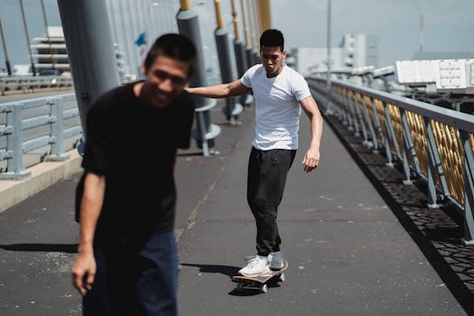 Young cheerful ethnic men riding skateboard on asphalt bridge with iron fence on cloudy day