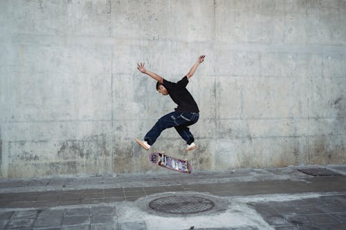 Young man jumping with skateboard above manhole near concrete wall