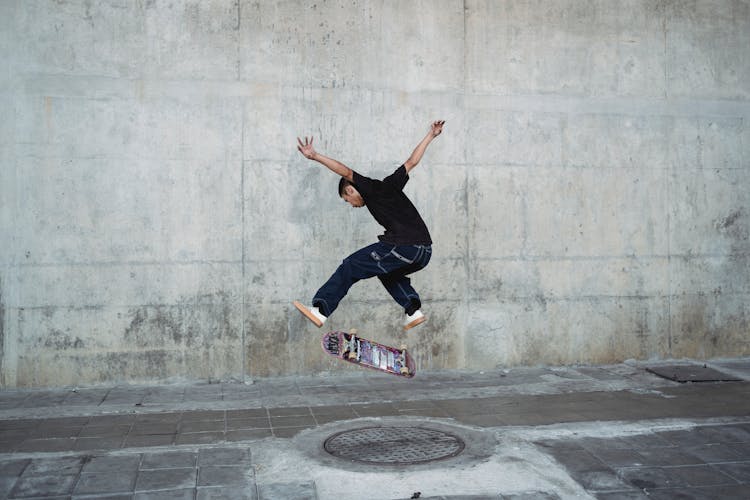 Young Man Jumping With Skateboard Above Manhole Near Concrete Wall