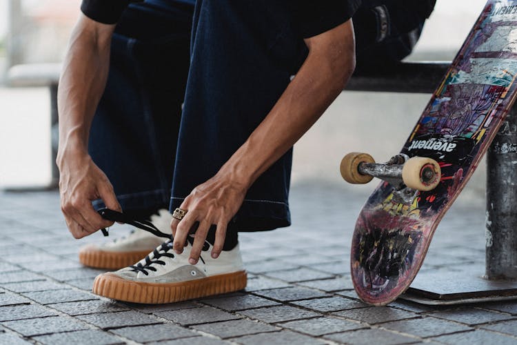 Crop Young Man Tying Shoelaces On White Sneakers Near Skateboard