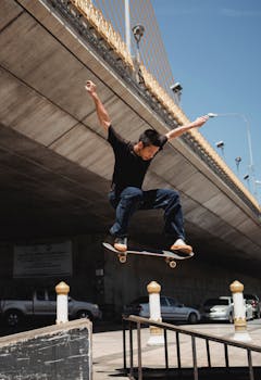 Sportive young Asian guy performing oliie stunt near staircase with railing on city street with high bridge and parked cars