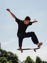 Full body of determined Asian guy jumping high on skateboard against blue sky in park on summer day