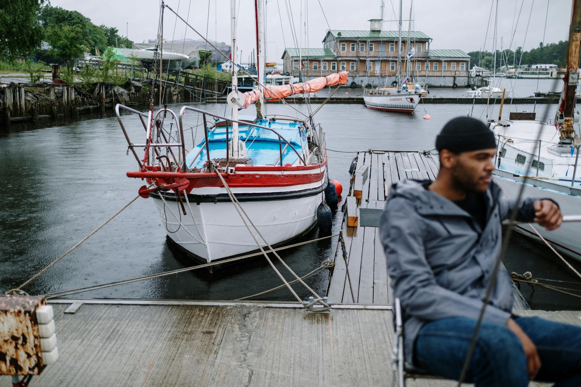 A man sits on a dock beside a sailboat in a harbor, cloudy skies overhead.