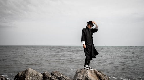 Free A Man in Black Outfit Standing on a Rock Near the Sea Stock Photo
