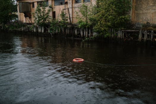 A lifebuoy floats in a rainy river by an abandoned building, creating a moody scene.