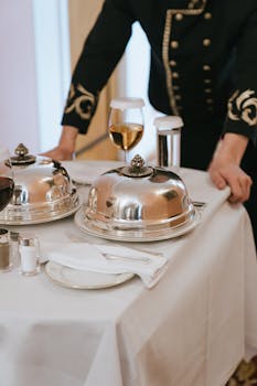 A classy dining scene featuring covered silver trays and a waiter preparing the table.