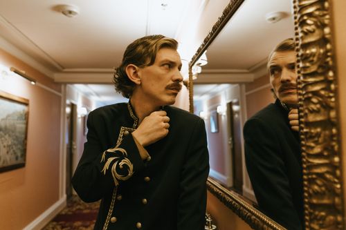 Free A bellboy in a luxurious hotel corridor adjusts his jacket, reflected in a large ornate mirror. Stock Photo