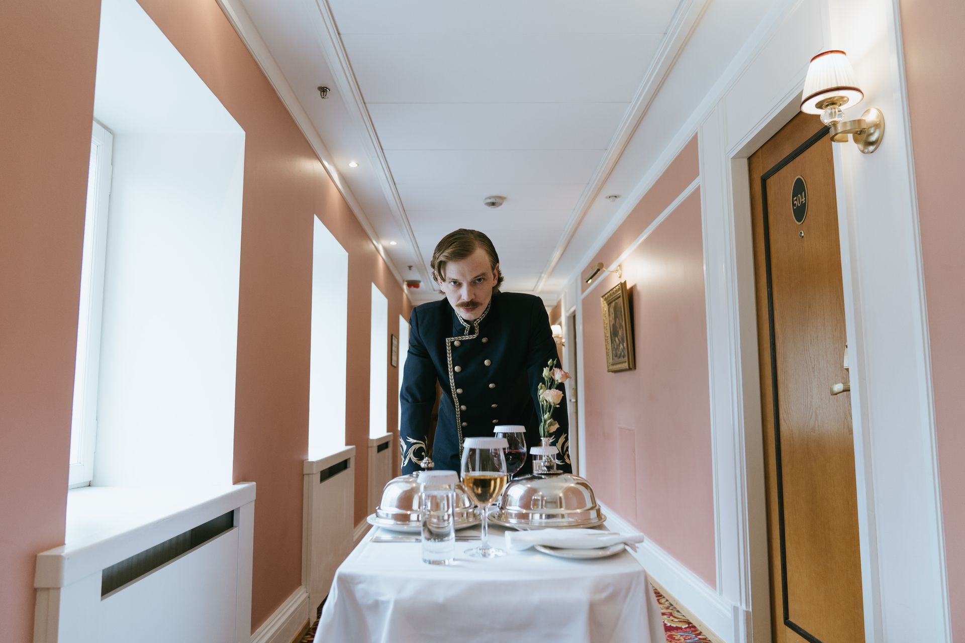 Bellboy in uniform presents room service in a hotel corridor setting.