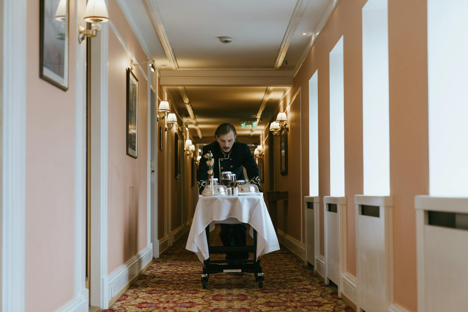 A hotel staff member pushes a room service trolley in a luxurious corridor.