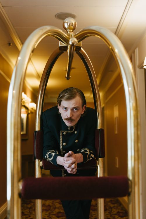 Free Bellboy with a mustache leaning on a hotel trolley in a hallway. Elegant and vintage style. Stock Photo