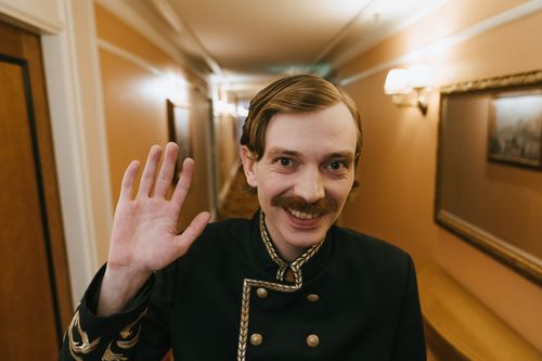 Free A smiling bellboy with a moustache waves in a hotel hallway, creating a welcoming atmosphere. Stock Photo