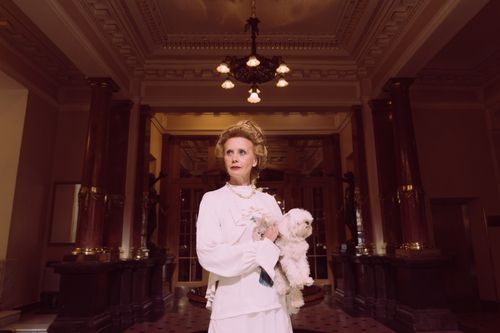 Free An elegant woman in a classic white dress holding a Maltese dog in a luxurious, vintage lobby. Stock Photo