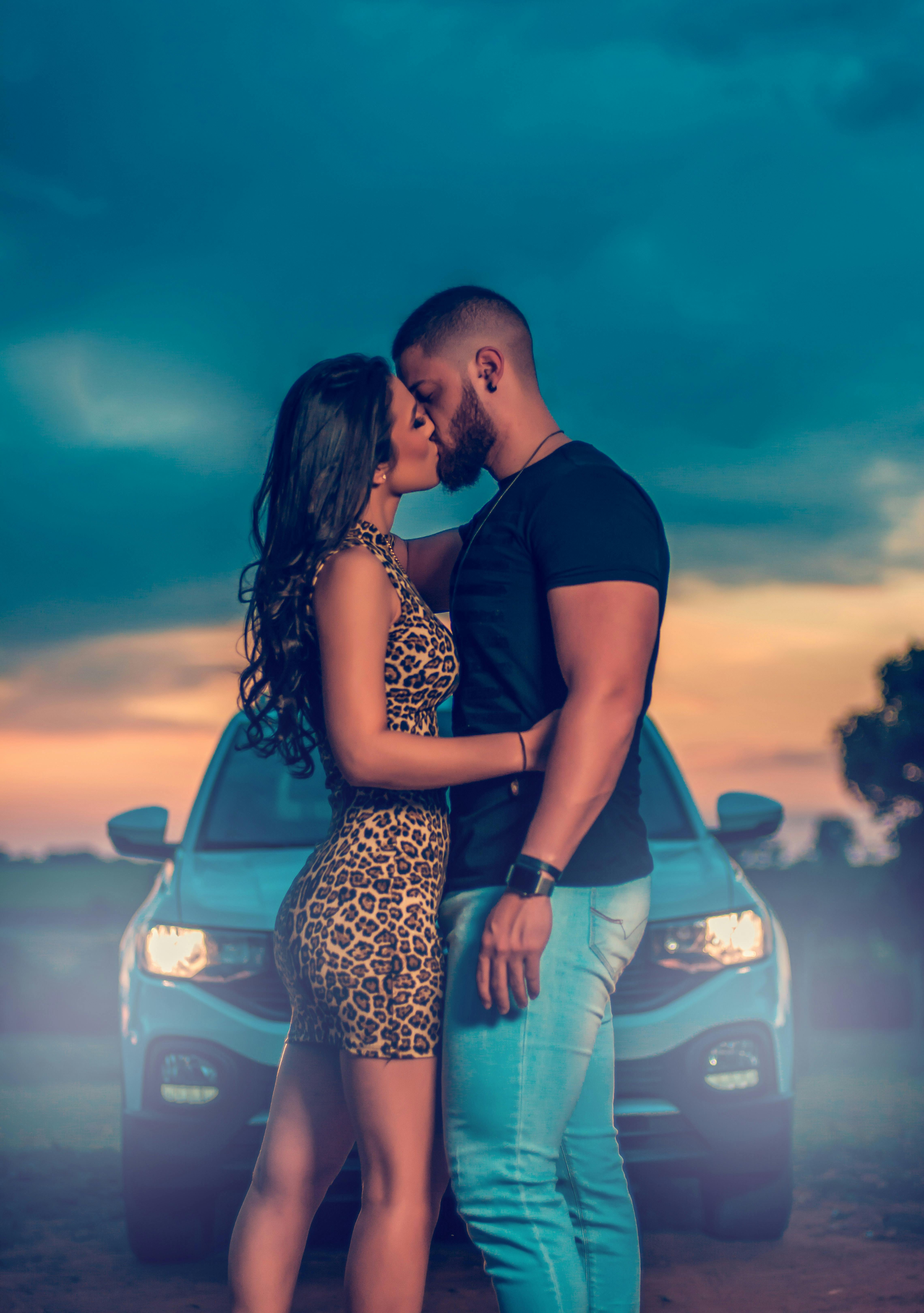 A couple kissing in front of a car during an overcast sunset, displaying a romantic moment.