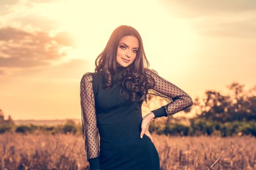 Free A beautiful woman in a black dress poses confidently in a sunlit field during sunset. Stock Photo
