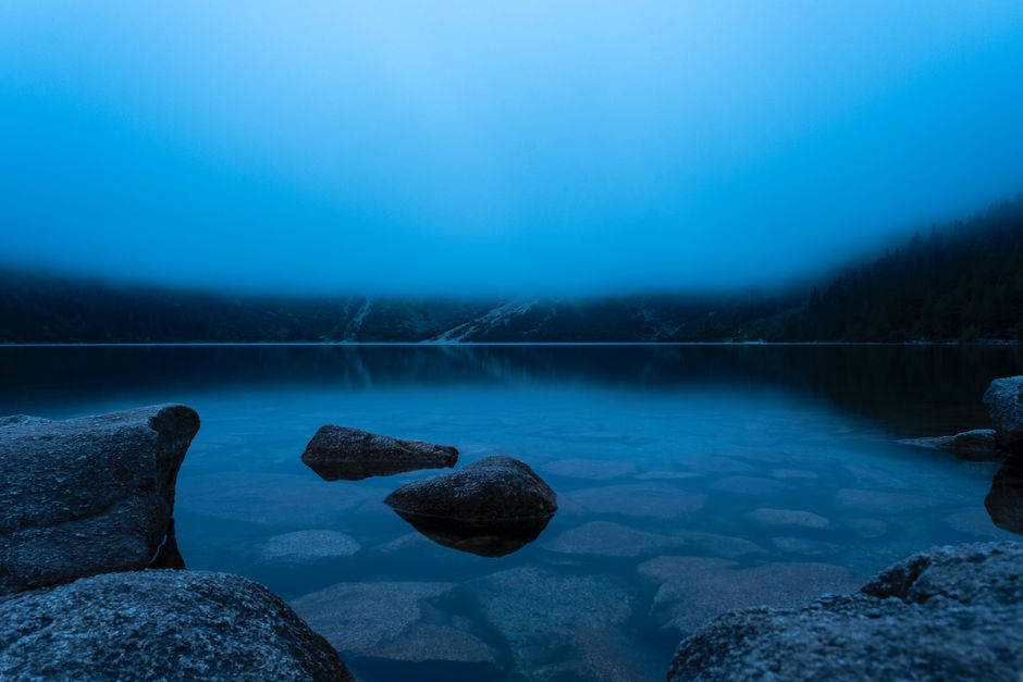 Calm mountain lake shrouded in fog, reflecting tranquil water and rocks.