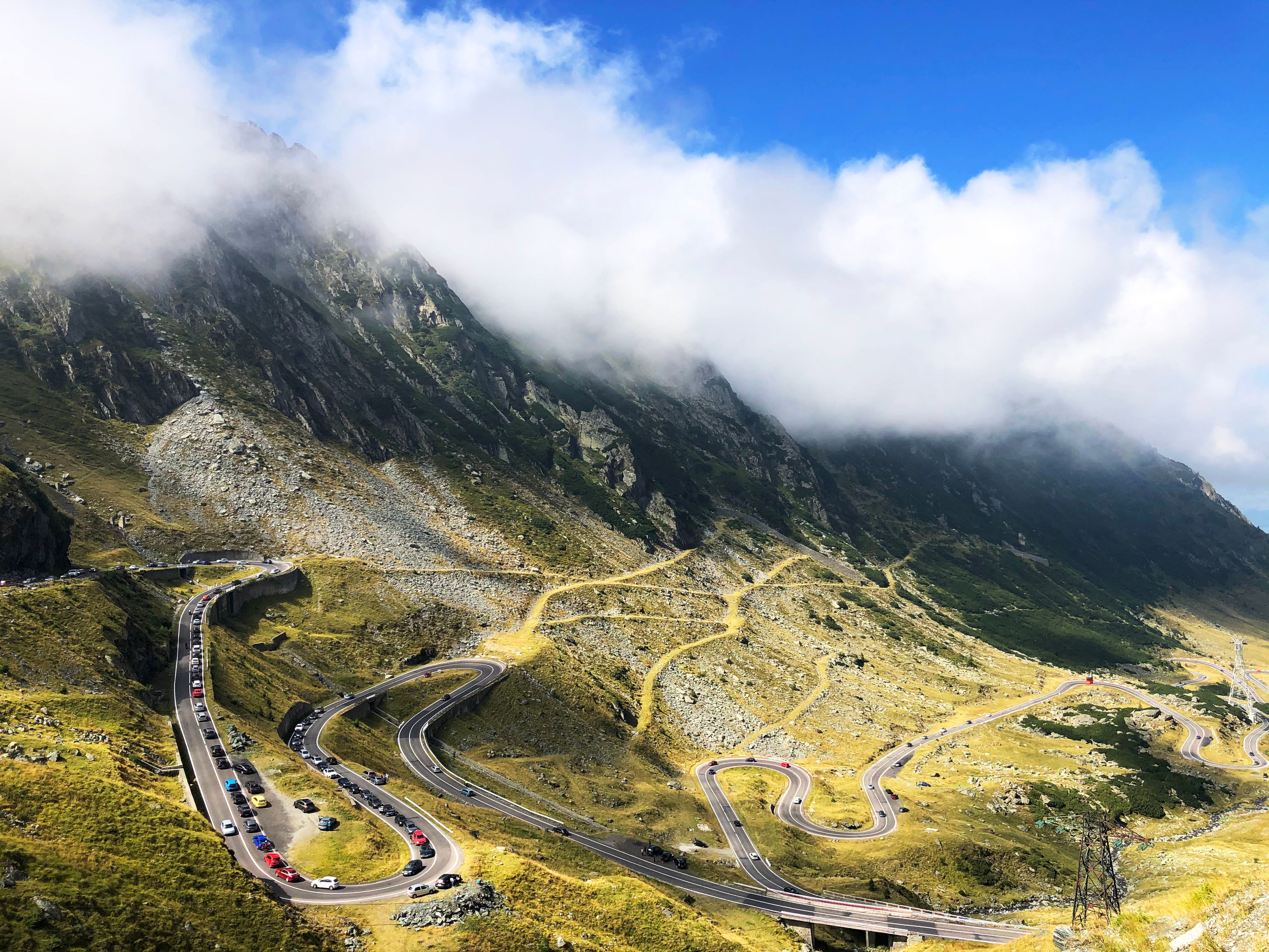 Winding mountain road curves through a highland landscape beneath low clouds and a bright blue sky