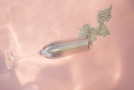 A champagne glass spilling silver glitter on a soft pink background, creating a sparkling artistic effect.