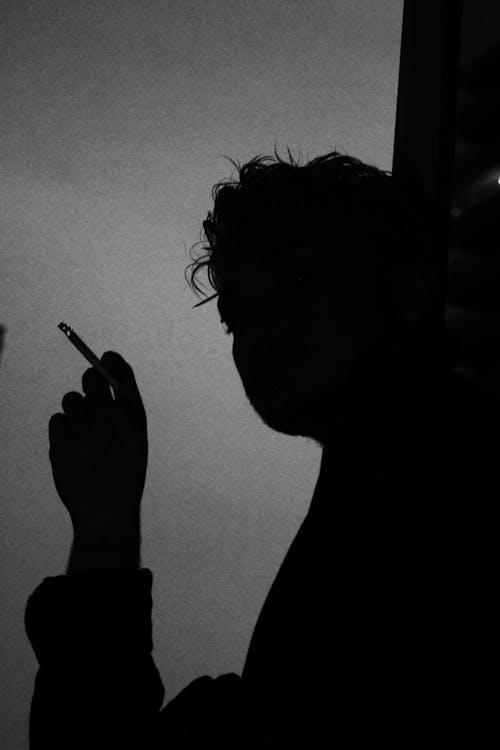 Black and white low angle side view of silhouette of person smoking cigarette while resting alone and thinking on problem
