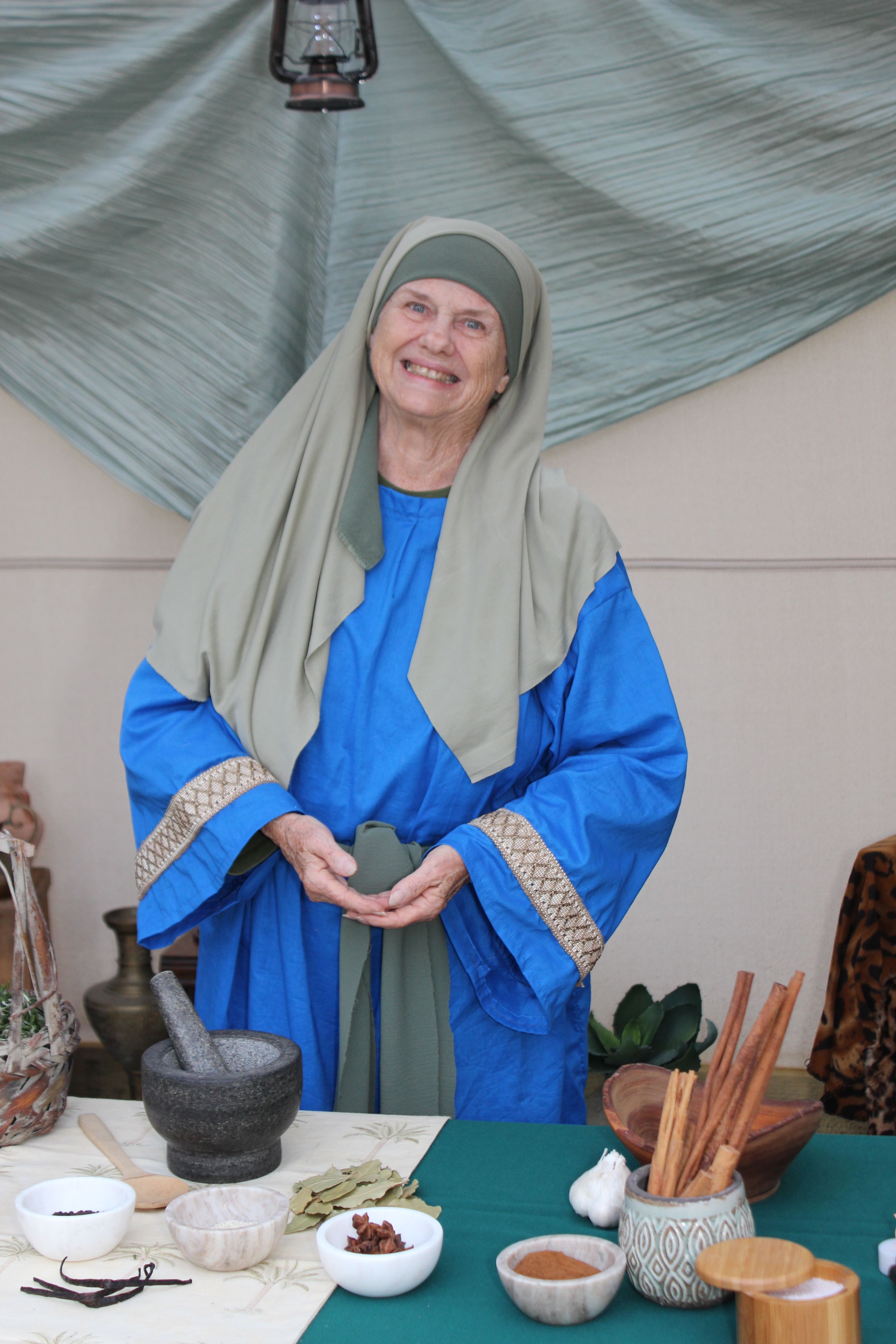 Smiling woman in traditional attire with herbal medicine setup indoors.
