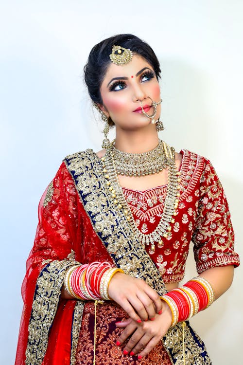 Dreamy young Indian female wearing traditional red clothes with makeup and bracelets with necklace and earrings with piercing looking away on white background