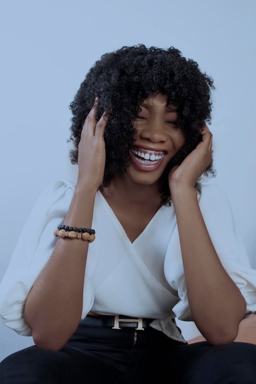 Young African American female keeping hands on head while laughing with closed eyes on white background