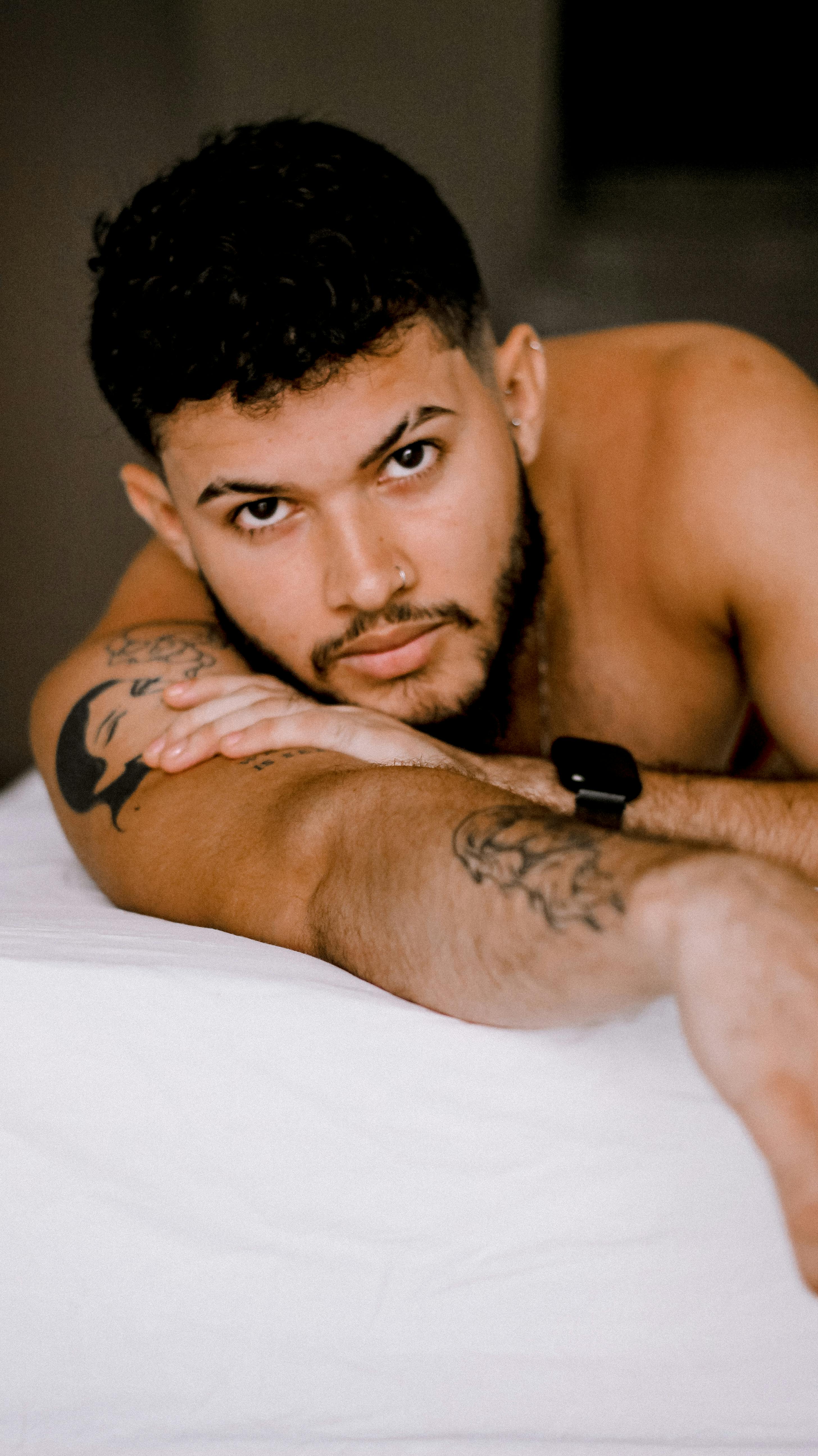 A tattooed man with a beard lying relaxed and topless on a bed indoors, gazing at the camera.