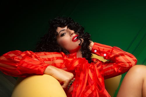 Low angle of confident sensual female with red lips touching hair sitting on chair and looking at camera