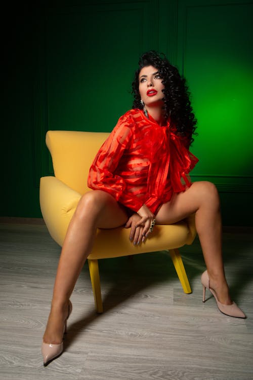 Full body of alluring female in blouse and high heels sitting on chair in studio and looking away