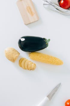 Flatlay of fresh eggplant, corn, and potato with knife on white surface for cooking preparation.