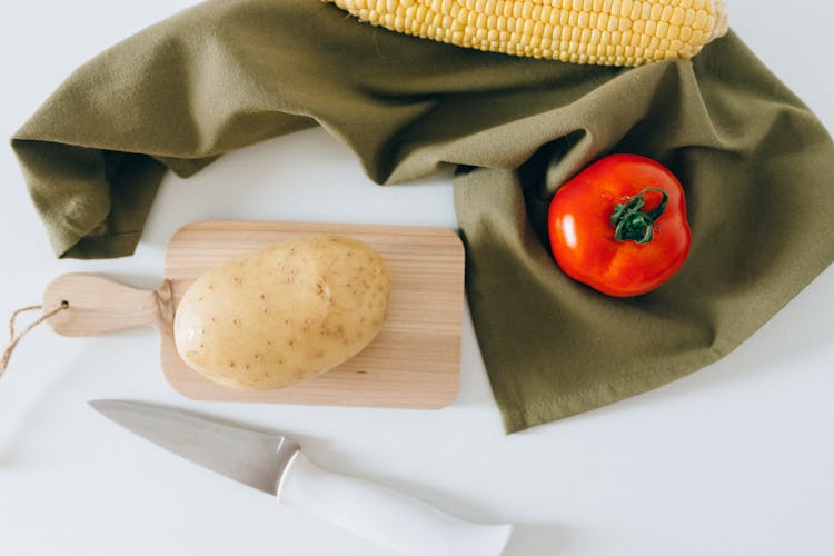 A Potato On A Wooden Chopping Board Beside A Tomato And Corn