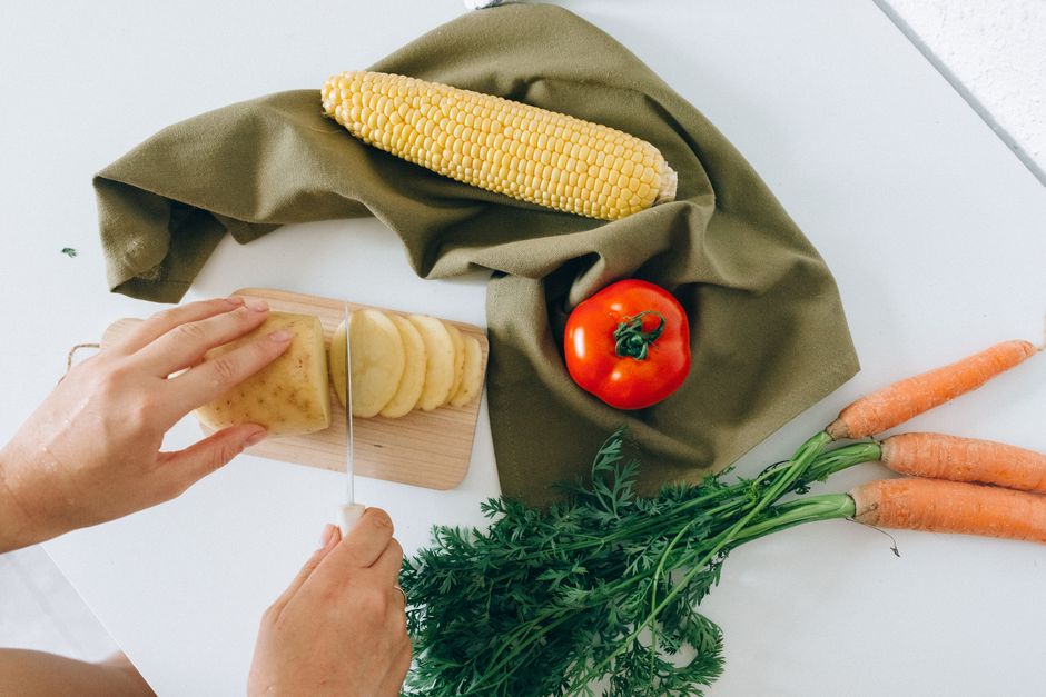 Photo by Nataliya Vaitkevich on Pexels Hands slicing potato with fresh vegetables like corn, tomato, and carrots on table. - Photo by Nataliya Vaitkevich on Pexels