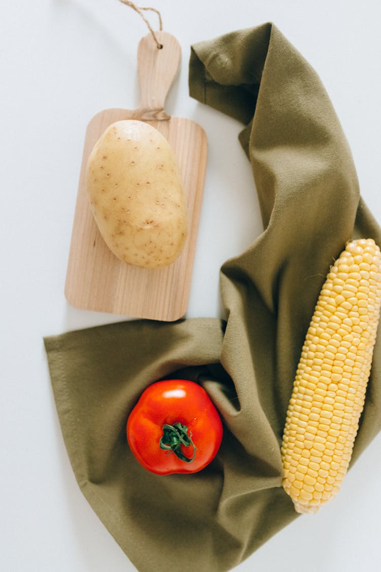 A Potato On A Wooden Chopping Board Beside A Tomato And Corn