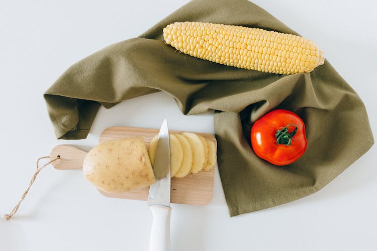 Slices Of Potato On A Wooden Chopping Board Beside A Tomato And Corn
