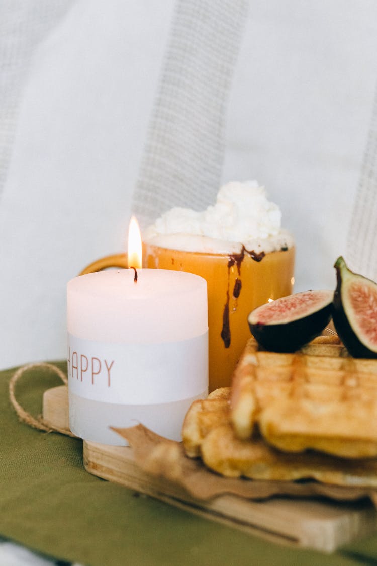 A Lighted Candle On A Wooden Chopping Board Beside A Mug With Cream
