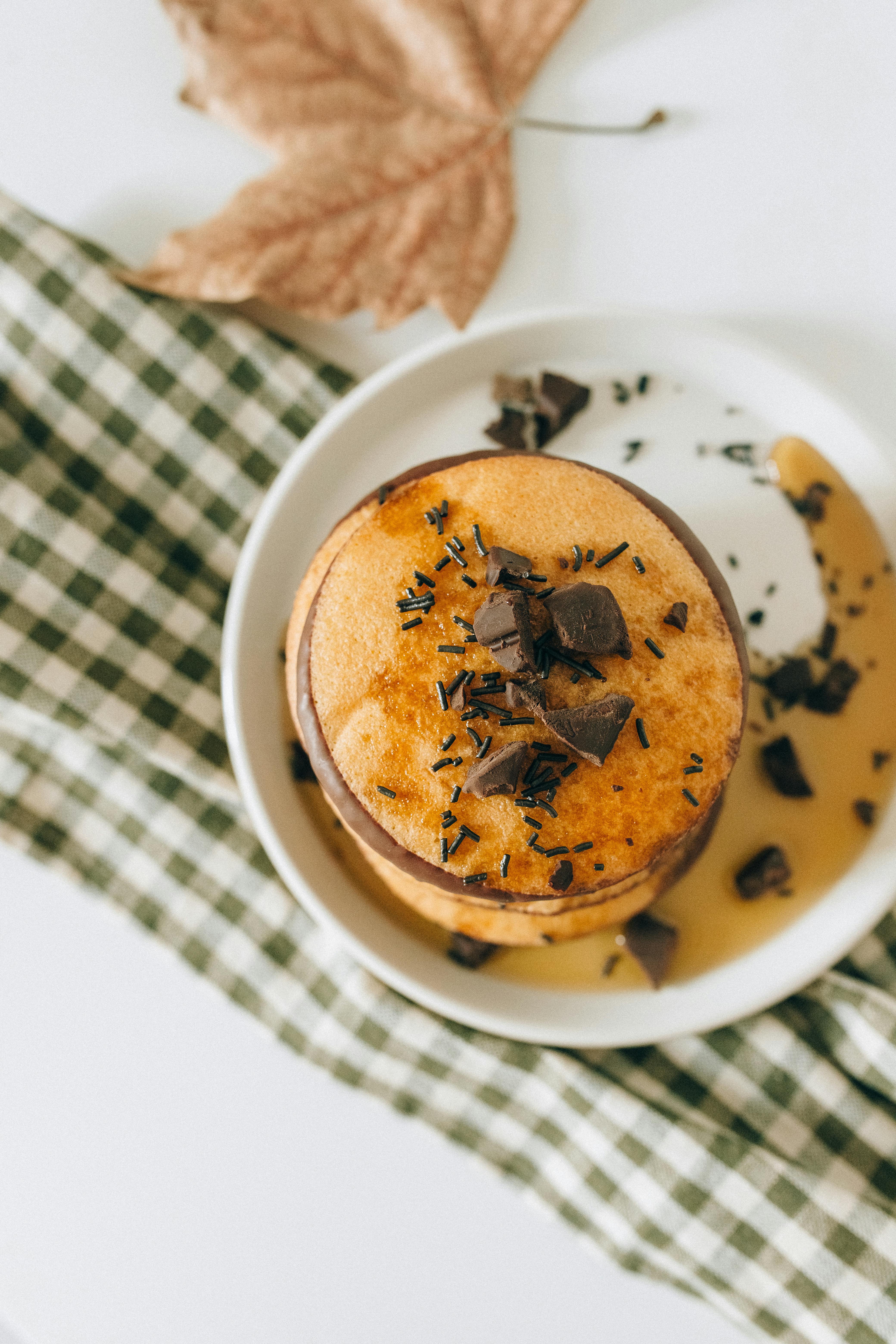 Aerial view of stacked pancakes with chocolate syrup and maple leaf on a plaid cloth, evoking autumn flavors.