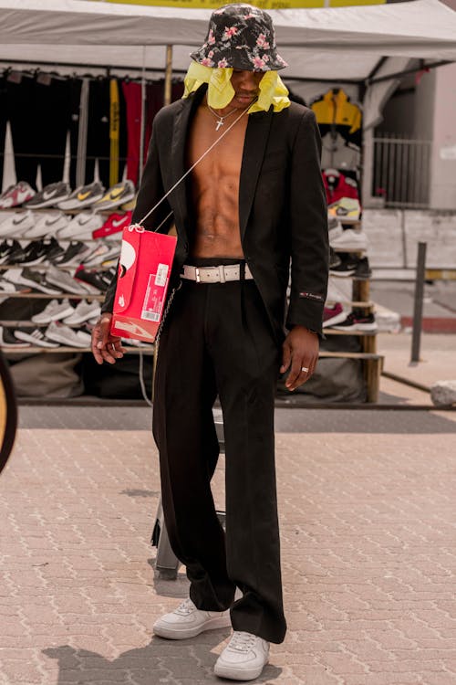 Full body of stylish African American male showing bare abs while standing near street shoe market