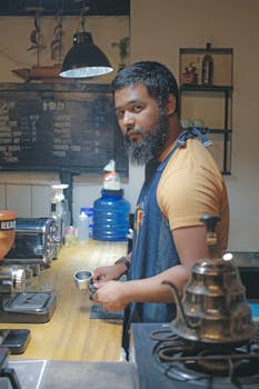 Barista with facial hair making coffee in an intimate café atmosphere, showcasing brewing equipment.