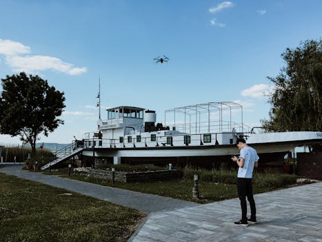 A man flying a drone near a historic boat in Hungary under a clear blue sky.