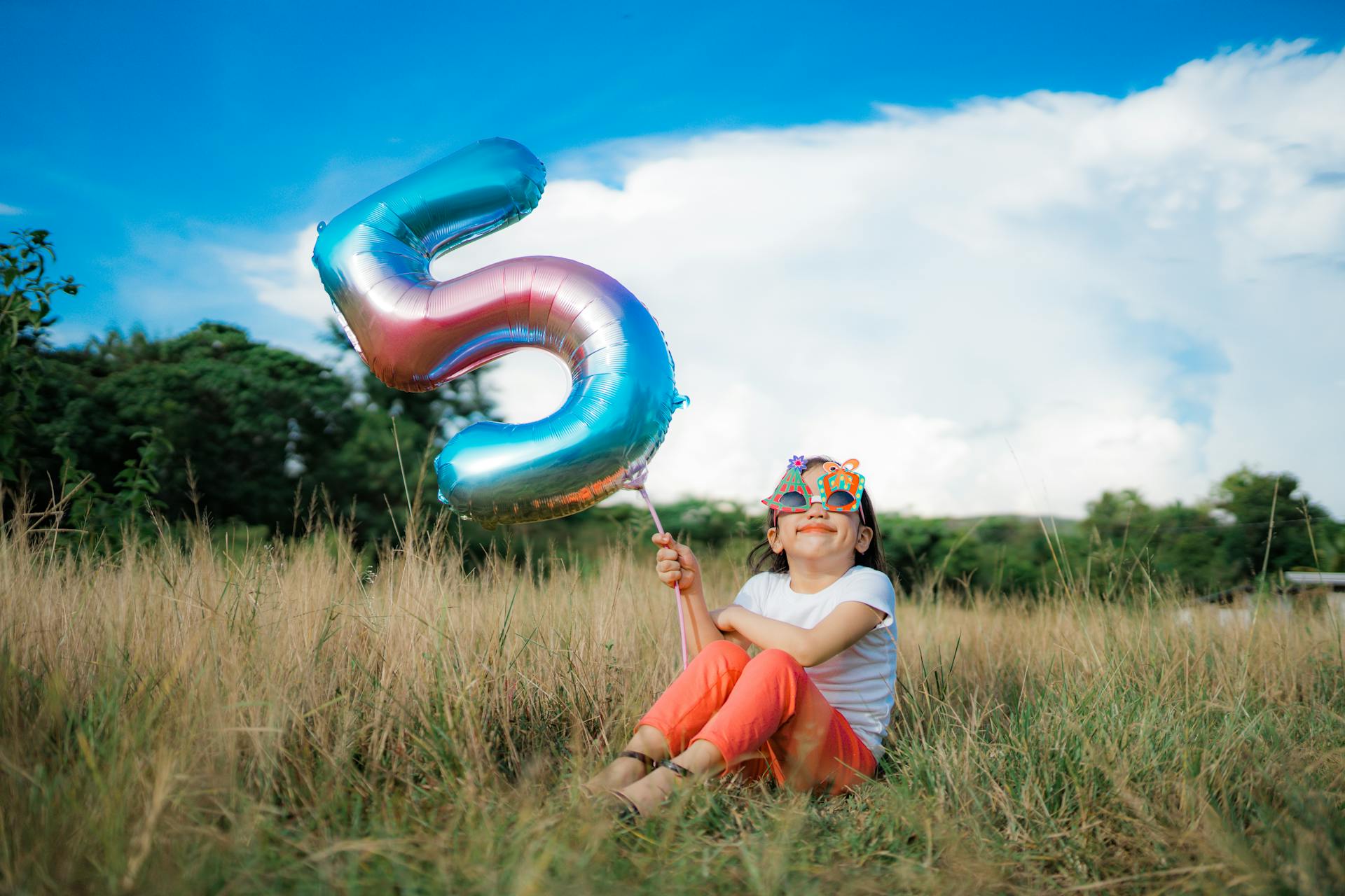 Young girl sitting outdoors holding a colorful number five balloon, wearing fun eyewear, enjoying a sunny day.